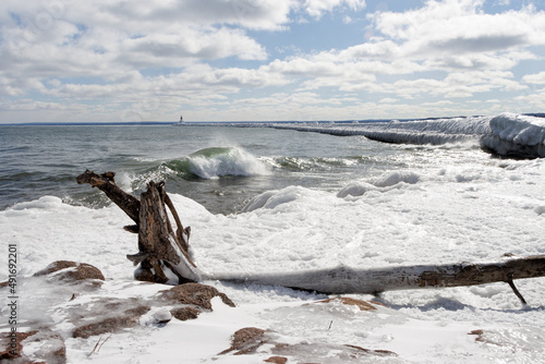 Waves Icy Lighthouse and Breakwall on Lake Superior in Marquette Michigan with Driftwood and Sandstone on a Winter Day with Puffy Blue Clouds in the Sky 