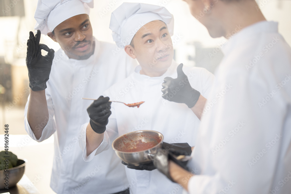 Three chef cooks with different ethnicities tasting sauce with a spoon while cooking in the kitchen. Teamwork and cooking delicious food at restaurant Three chef cooks with different ethnicities tasting sauce with a spoon while cooking in the kitchen. Teamwork and cooking delicious food at restaurant