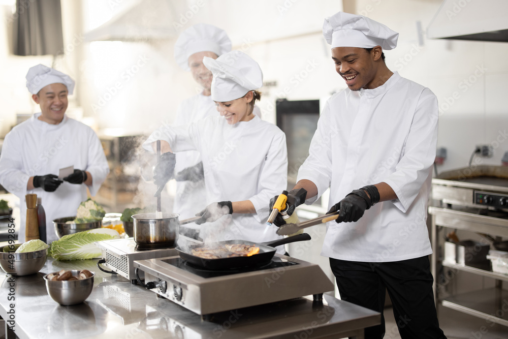 Multiracial team of professional cooks in uniform preparing meals for a restaurant in the kitchen. Latin guy frying meat, european cooks making sauce and asian chef managing the process. Teamwork and Multiracial team of professional cooks in uniform preparing meals for a restaurant in the kitchen. Latin guy frying meat, european cooks making sauce and asian chef managing the process. Teamwork and