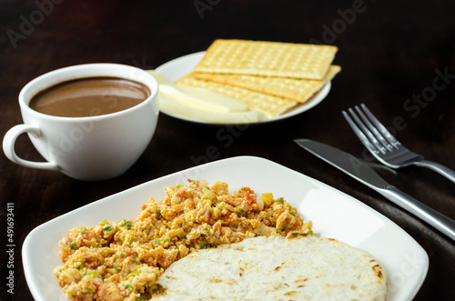 Colombian breakfast.Arepa (ground corn flour dough),egg with traditional sauce called hogado(stir-fried tomato and onion),with cup of chocolate,crackers and cheese,on background dark wood. Close up.