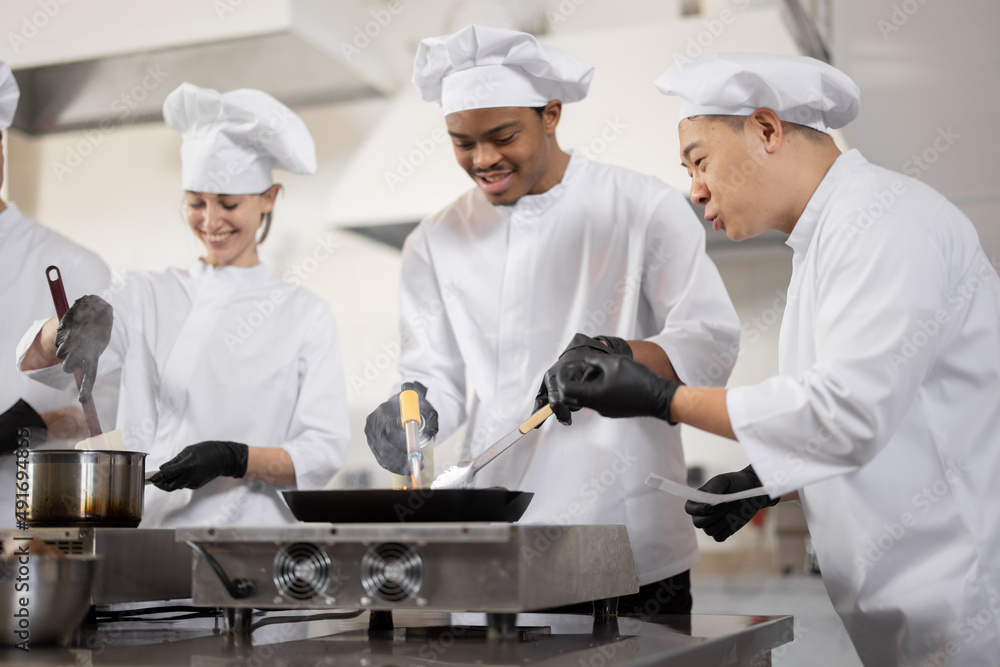 Multiracial team of professional cooks in uniform preparing meals for a restaurant in the kitchen. Asian chef managing the process, latin guy and european cooks frying. Teamwork and hard job at