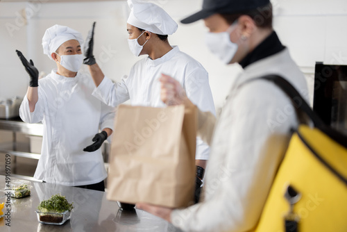 Courier waiting for an order for delivery in the kitchen with chefs preparing takeaway food. Concept of dark kitchen and delivering food during pandemic. Multiracial cook team in face mask and