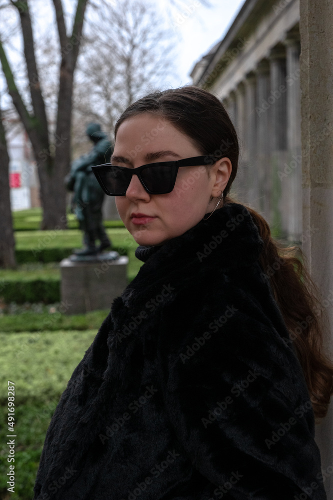Portrait of a young woman in all-black outfit and black sunglasses on a grey cloudy day in park. Black clothes, typical Berlin. Green cut bushes, statue, columns