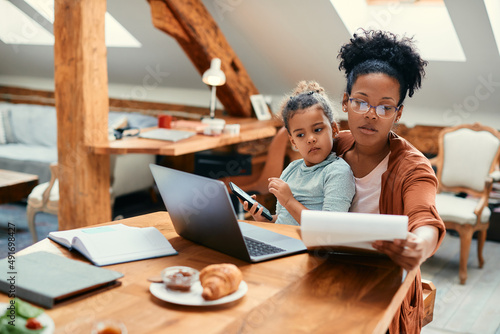 African American working mother using laptop and analyzing paperwork while daughter is sitting on her lap at home.