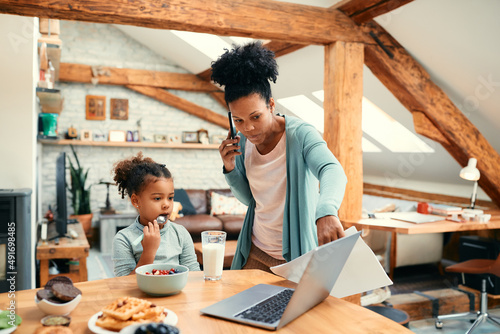 Black little girl eats while busy mother is giving her laptop to use during breakfast. photo