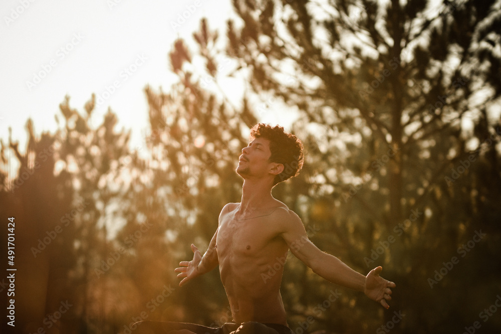 Young man doing yoga at the sunrise on a top of a mountain next the trees over the rocks with arm wide open Young man doing yoga at the sunrise on a top of a mountain next the trees over the rocks with arm wide open