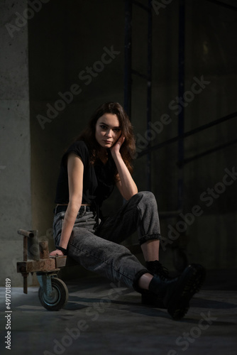 A beautiful young girl sits on scaffolding in a beam of light. Dressed in jeans and black boots
