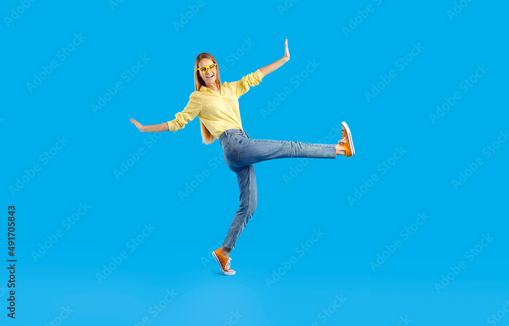 Funny cheerful pretty girl in comfortable casual clothes having fun in studio. Happy slim young woman in shirt, jeans, eyeglasses and canvas sneakers walking isolated on solid blue background