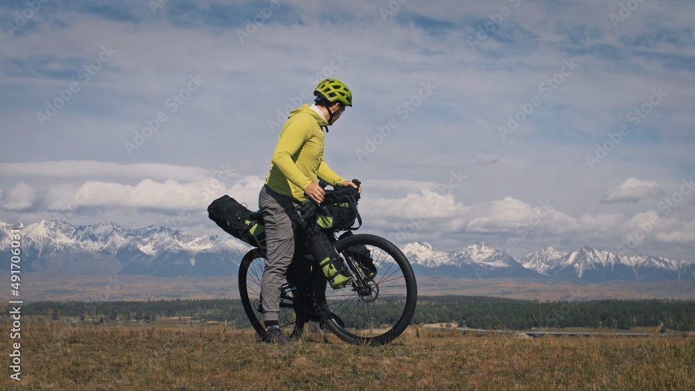The man travel on mixed terrain cycle touring with bikepacking. The traveler journey with bicycle bags. Sport tourism bikepacking, bike, sportswear in green black colors. Mountain snow capped. The man travel on mixed terrain cycle touring with bikepacking. The traveler journey with bicycle bags. Sport tourism bikepacking, bike, sportswear in green black colors. Mountain snow capped.