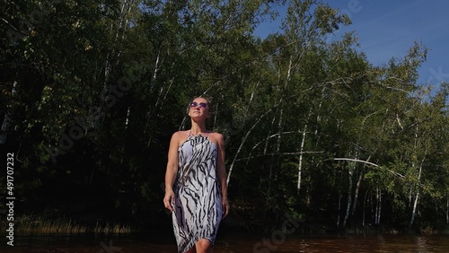 Woman walk on water on pier in sunglasses and a boho silk shawl. Girl rest on a flood wood underwater dock. The pavement is covered with water in the lake. In the background are mountain and a forest.