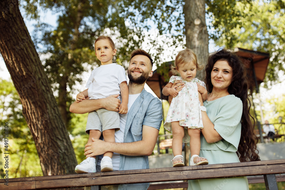 Fototapeta premium Lovely parents with little kids at the park, caring father hold in arms joyful son, cute daughter sitting in mothers hands, enjoy happy family moments, weekend outdoors concept