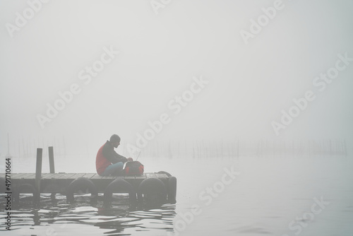 Digital nomad travelling while working with laptop on wooden dock by the sea surrounded by mist