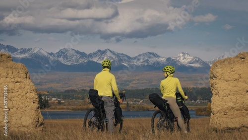 The man and woman travel on mixed terrain cycle bike touring with bikepacking. The two people journey with bicycle bags. Sport sportswear in green black colors. Mountain snow capped, stone arch. photo