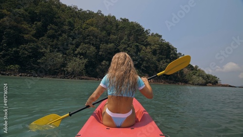 Pretty sexy young blonde woman in stylish swimsuit enjoys trip canoe on sea against palm islands. Traveling to tropical countries. Positive sports girl hand padding on kayak, back view. © ivandanru