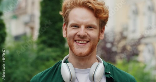 Portrait of Healthy Smiling Ginger Guy standing in Park looking at the Camera. Having Nice Haircut and Headphones on neck. Spending Time outdoors. Having Attractive Appearance with Freckles.