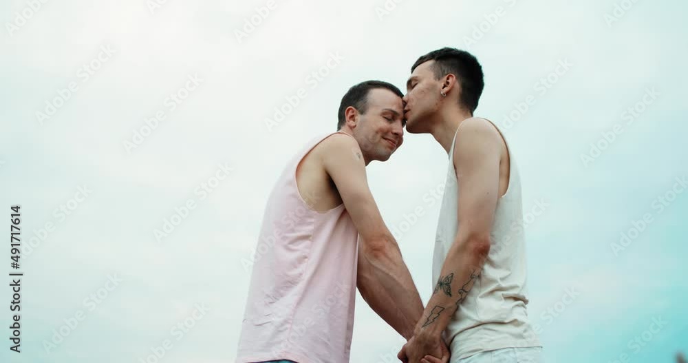 Portrait of Young LGBT Men Couple in Love Holding Hands while standing in a Field. Two Handsome Guys Lovers looking at Each Other with Beautiful Blue Sky Background. Equality Rights of People. Portrait of Young LGBT Men Couple in Love Holding Hands while standing in a Field. Two Handsome Guys Lovers looking at Each Other with Beautiful Blue Sky Background. Equality Rights of People.