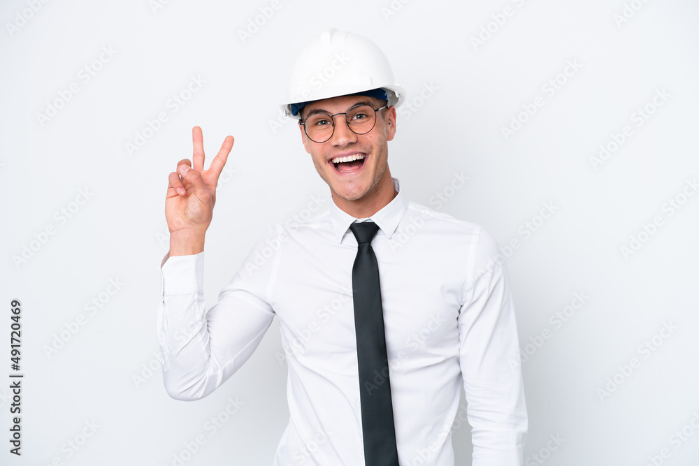 Young architect caucasian man with helmet and holding blueprints isolated on white background smiling and showing victory sign