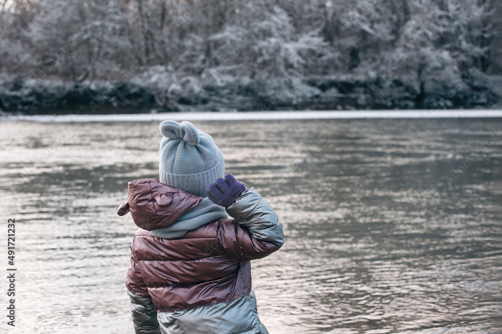 a little girl on a winter walk stands with her back to us and throws stones into a cold river a little girl on a winter walk stands with her back to us and throws stones into a cold river