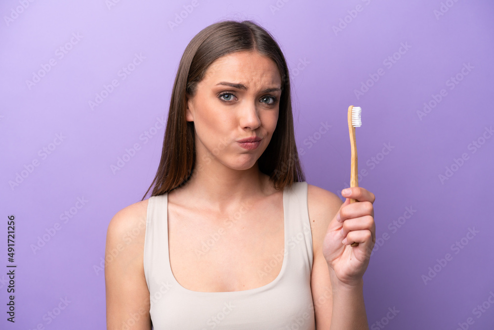 Obraz premium Young caucasian woman brushing teeth isolated on purple background with sad expression