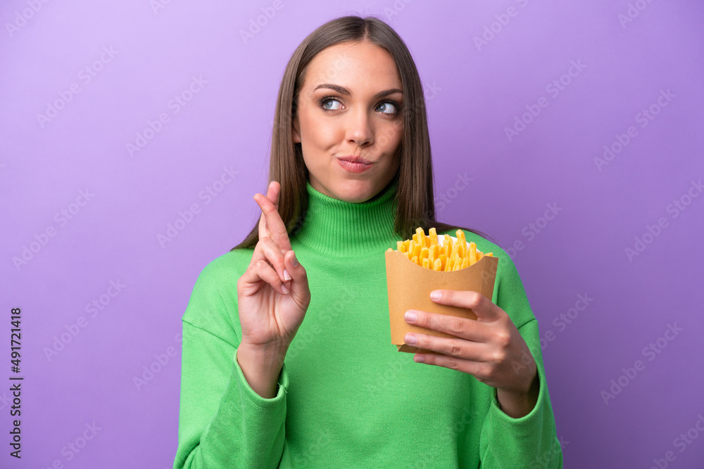 Young caucasian woman holding fried chips on purple background with fingers crossing and wishing the best Young caucasian woman holding fried chips on purple background with fingers crossing and wishing the best
