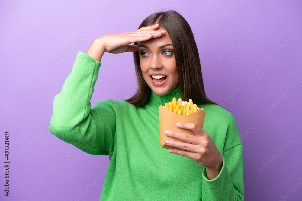Young caucasian woman holding fried chips on purple background doing surprise gesture while looking to the side Young caucasian woman holding fried chips on purple background doing surprise gesture while looking to the side
