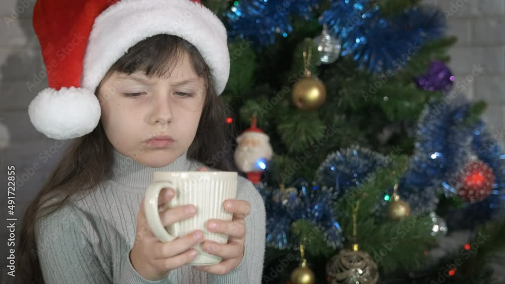 Get warm with tea by christmas tree. A little girl in Santa hat get warm with a hot mug of tea in loneliness during Christmas holidays. Get warm with tea by christmas tree. A little girl in Santa hat get warm with a hot mug of tea in loneliness during Christmas holidays.