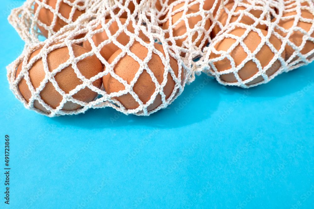 Naklejka premium Natural brown hen eggs in white mesh bag on blue background. Creative minimal abstract Easter holiday concept.