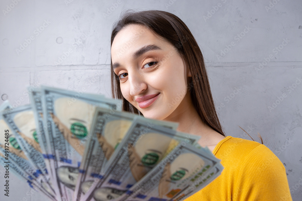 Photo of a wealthy woman in simple clothes holding a fan of dollar money isolated against a concrete wall background Photo of a wealthy woman in simple clothes holding a fan of dollar money isolated against a concrete wall background