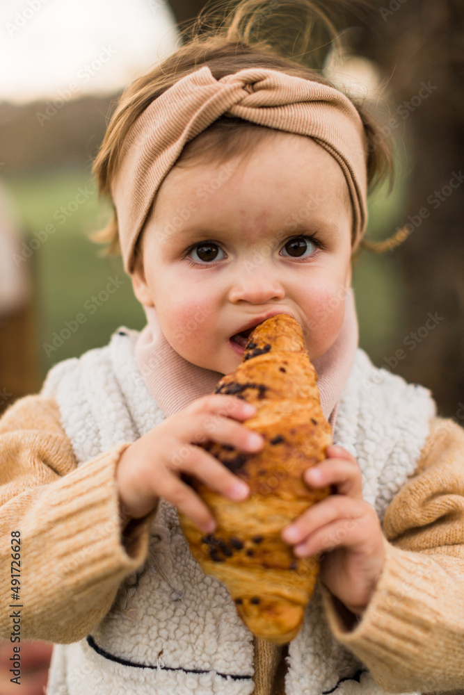 Little girl in beige coat eating croissant. Looking at front,to the camera.Autumn or spring photo. Little girl in beige coat eating croissant. Looking at front,to the camera.Autumn or spring photo.