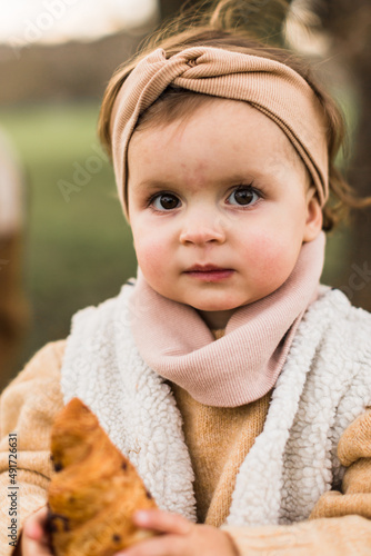 Little girl in beige coat eating croissant. Looking at front,to the camera.Autumn or spring photo.
