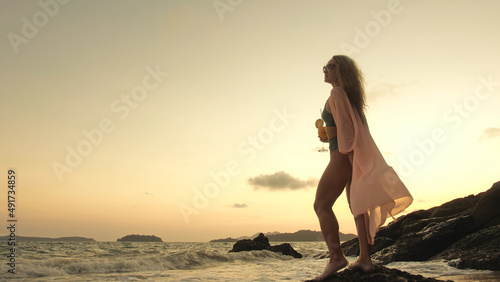 Silhouette woman stand on a reef rock stone in sea on golden sunset. Girl on tropical beach in green swimsuit and flutter in wind pink tunic silk shirt cape, drinks her orange cocktail Pina Colada