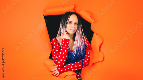 Smiling woman with Senegalese braids in a hole in the wall. A stylish girl in a bright red dress looks at the camera through a hole in the orange wall in the studio