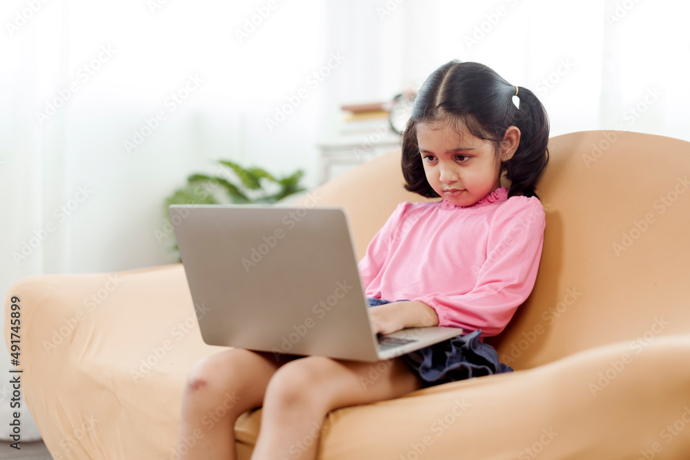 An adorable Asian girl wearing a pink shirt is happily sitting at her laptop notebook computer on the sofa in the living room of her home. An adorable Asian girl wearing a pink shirt is happily sitting at her laptop notebook computer on the sofa in the living room of her home.