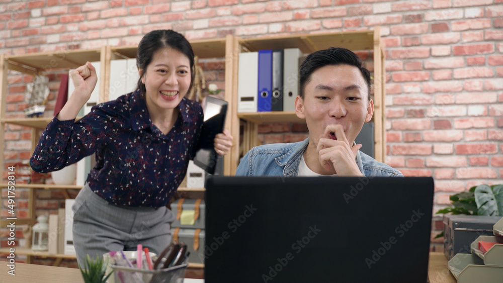 asian female is covering her mouth in surprise and holding fist up to cheer as her startups business team partner using laptop is gesturing to check out the news. asian female is covering her mouth in surprise and holding fist up to cheer as her startups business team partner using laptop is gesturing to check out the news.