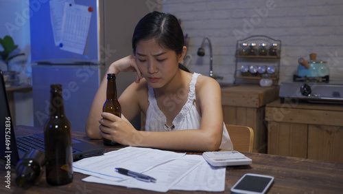 asian female seated at table with banking bills and a computer is drinking alcohol to release the pressure of her poor personal finance at home.