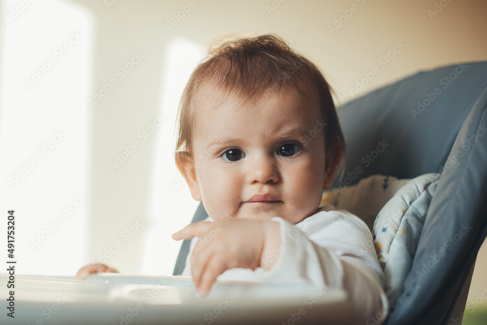 Caucasian baby girl sitting in high chair and looking curious into the camera. Baby care. Morning meal. Caucasian baby girl sitting in high chair and looking curious into the camera. Baby care. Morning meal.