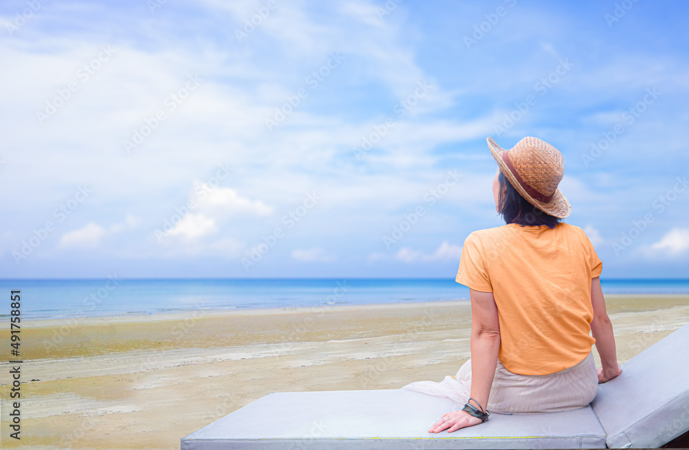 A young woman sits on a bench on the beach, looking at the sea. Concept image of relaxing by the sea, bright sky, white clouds on the beach.