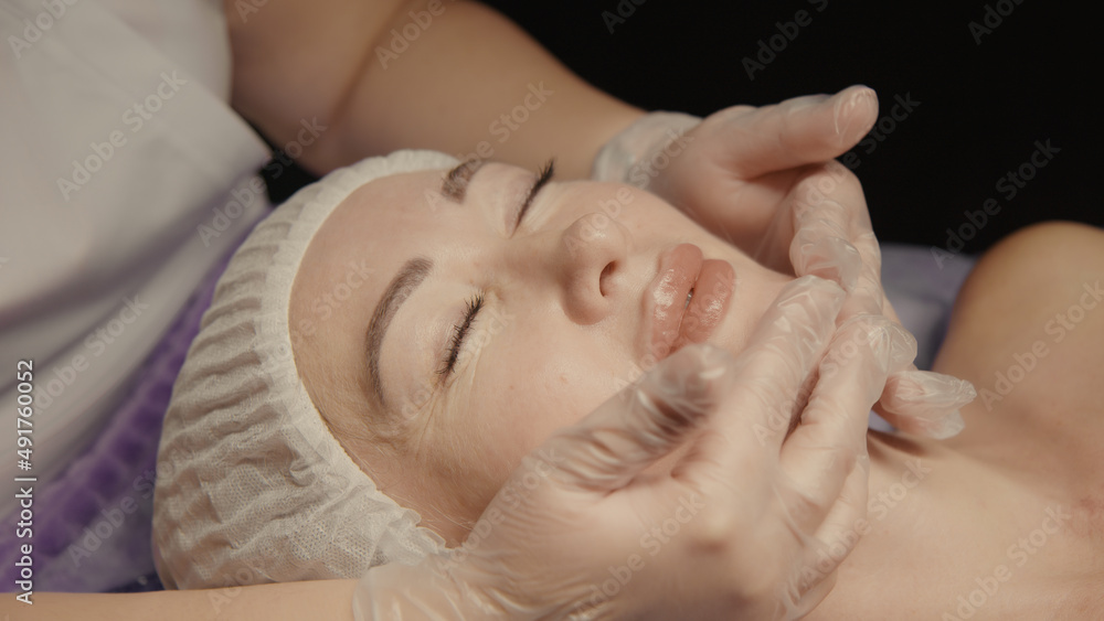Young hypnotic face woman receiving anti-ageing facial massage in spa salon relax. Wellness body skin care face beauty treatment, rejuvenation procedure getting head face massage. Black background Young hypnotic face woman receiving anti-ageing facial massage in spa salon relax. Wellness body skin care face beauty treatment, rejuvenation procedure getting head face massage. Black background