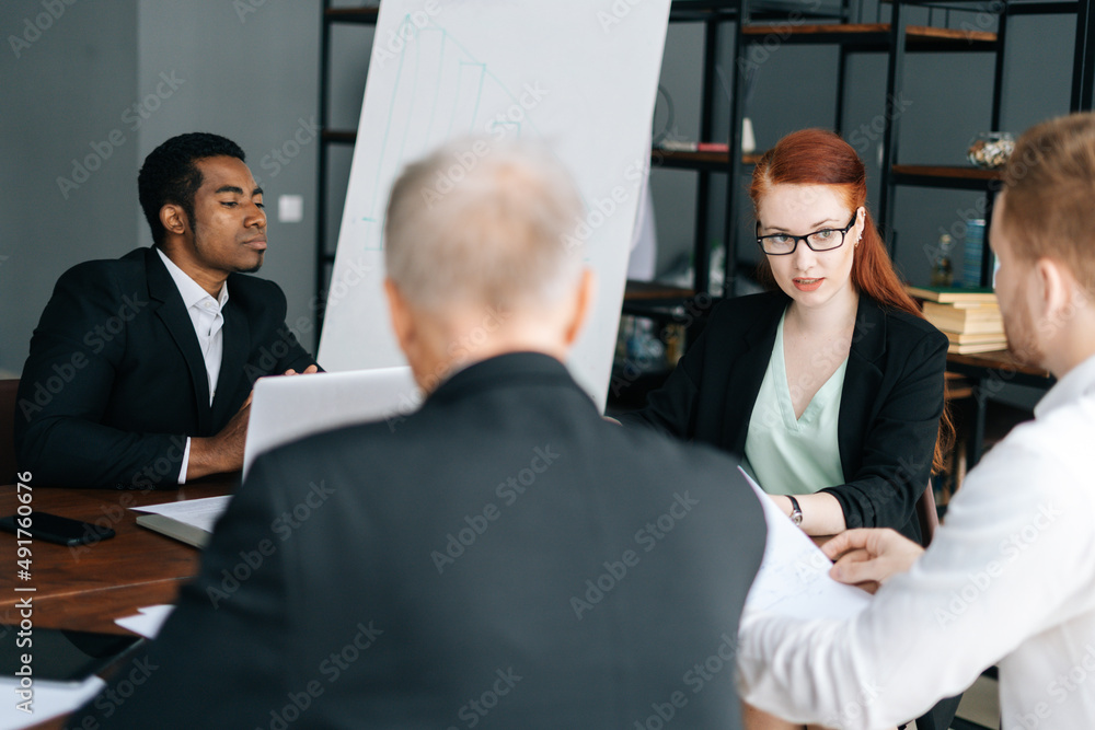 Focused female team leader in glasses holding corporate meeting with multi-ethnic colleagues in boardroom sitting at desk. Attractive businesswoman discussing project with business partners in office. Focused female team leader in glasses holding corporate meeting with multi-ethnic colleagues in boardroom sitting at desk. Attractive businesswoman discussing project with business partners in office.