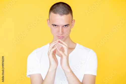 Sarcastic man. Evil look. Bad intention. Conspirator mockery guy in white t-shirt holding hands together showing sardonic smile isolated yellow. photo