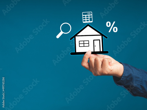 Hand holding a mini paper house with business icons while standing on a blue background. Business and real estate concept