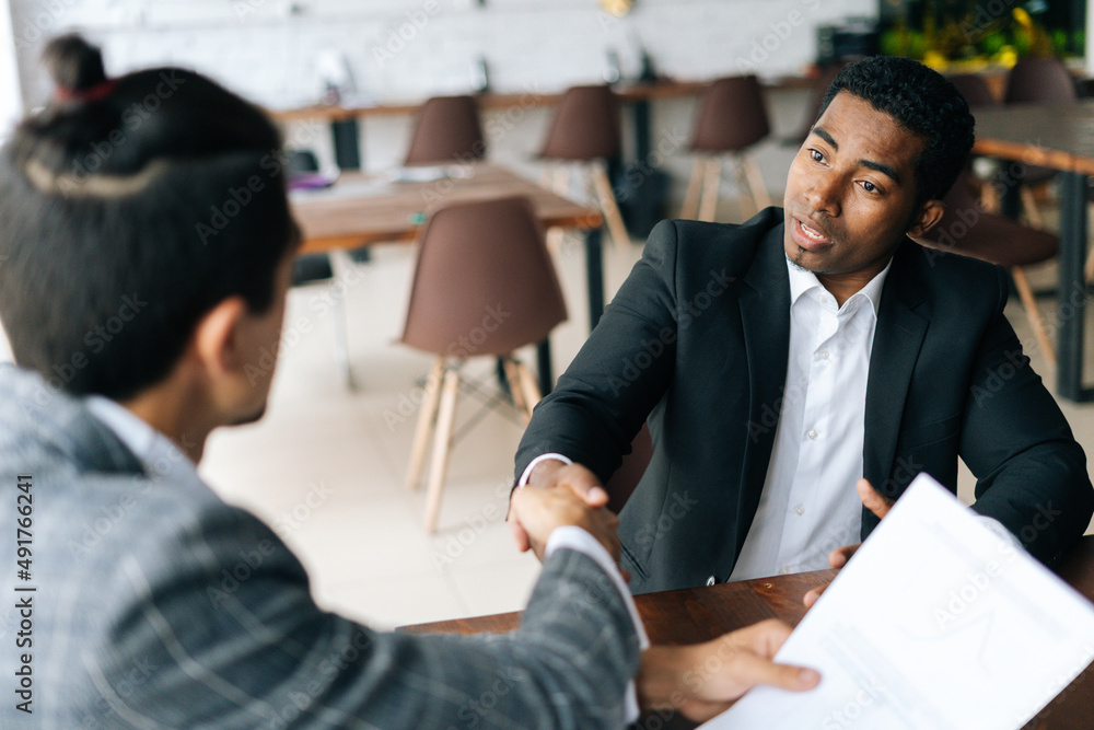 Close-up to two satisfied multiracial businessmen shaking hands over desk after successful negotiations, closing sealing deal, sitting at desk in modern office room. Concept of interracial cooperation Close-up to two satisfied multiracial businessmen shaking hands over desk after successful negotiations, closing sealing deal, sitting at desk in modern office room. Concept of interracial cooperation