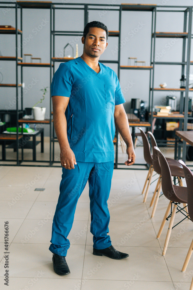 Vertical portrait of confident African American male doctor wearing blue surgeon medical uniform standing in hospital office, looking at camera. Front view of serious practitioner posing at workplace.