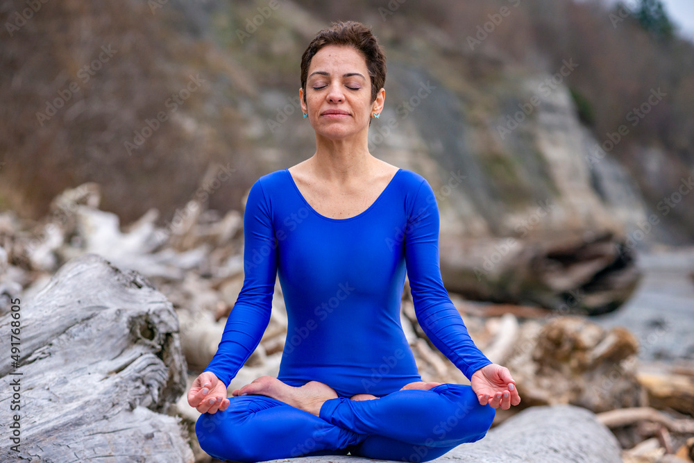 Athletic woman sitting in a yoga pose on driftwood with her eyes closed in deep meditation on a rugged Pacific Northwest beach.