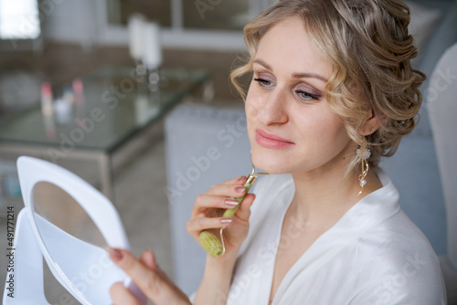 Cute caucasian woman is doing an anti-wrinkle facial massage with special roller holding mirror in her hands in silk bathrobe while sitting in an armchair in living room. Medicine beauty procedures