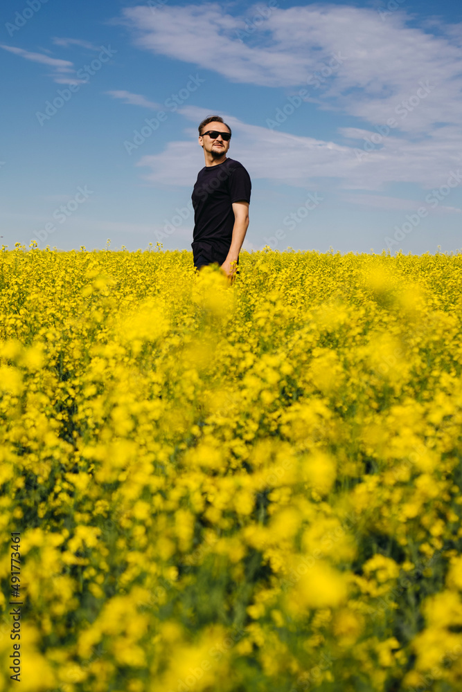 Man in black t-shirt is enjoying nature in the middle of field full of yellow flowers. Yellow field of flowering rapeseed with cloudy blue sky - brassica napus - plant for green energy, medicine.