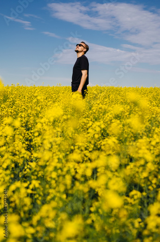 Man in black t-shirt is enjoying nature in the middle of field full of yellow flowers. Yellow field of flowering rapeseed with cloudy blue sky - brassica napus - plant for green energy, medicine. © Margarita Timofeeva