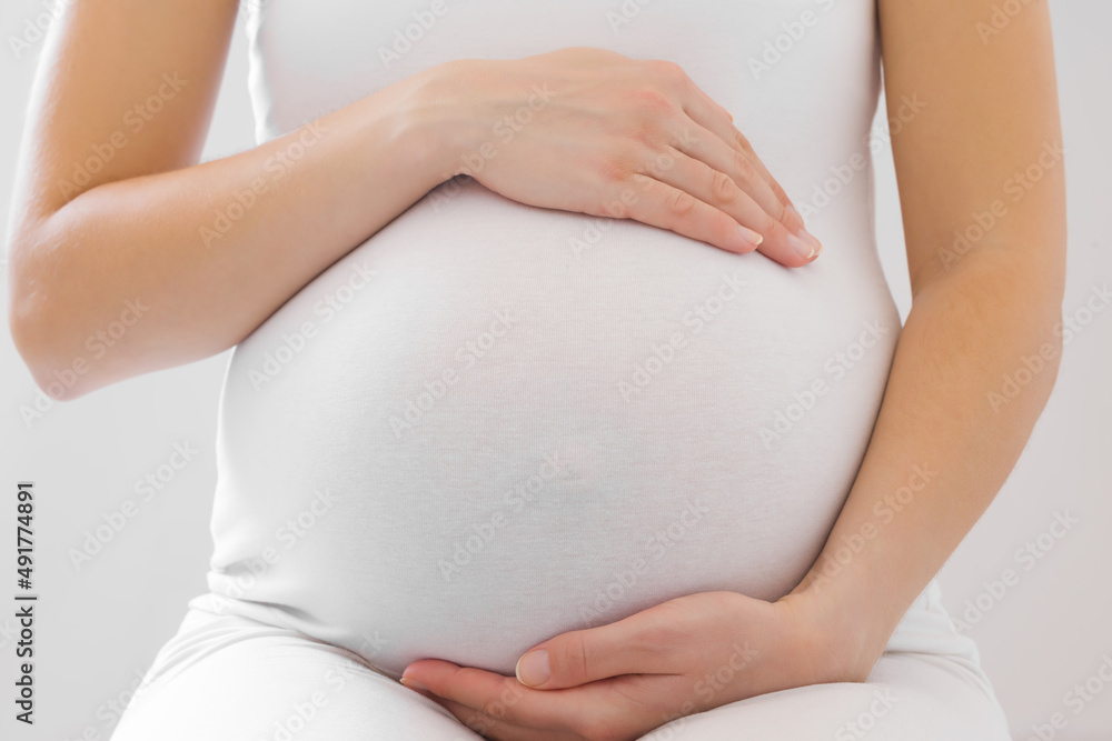Young adult pregnant woman in white clothes touching big belly and feeling her baby move on light gray background. Pregnancy time concept. Expectation time. Closeup. Front view. - obrazy, fototapety, plakaty 