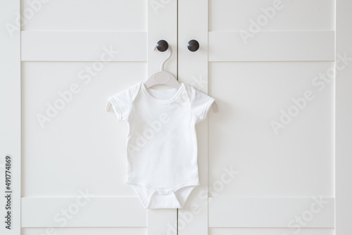 White baby bodysuit hanging on hanger at door of wardrobe. Closeup. Front view. Clothes preparing for newborn. photo