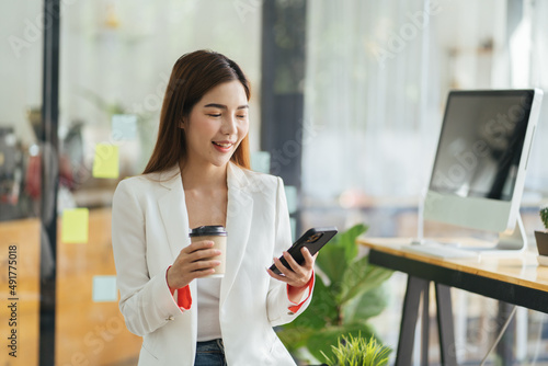 Asian business woman in cafe using mobile phone. Girl chatting on smartphone and drinking coffee in restaurant near window, smiling at cellphone screen.